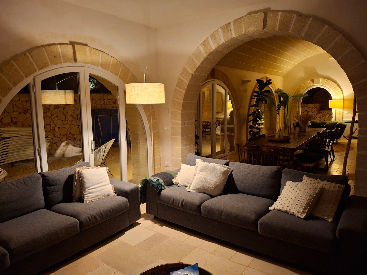 An inviting living area features two soft gray sofas adorned with decorative cushions. The stone archways lead to a well-lit dining area, showcasing a wooden table surrounded by chairs. Soft lighting creates a relaxed ambiance, enhancing the connection to the outdoor space.