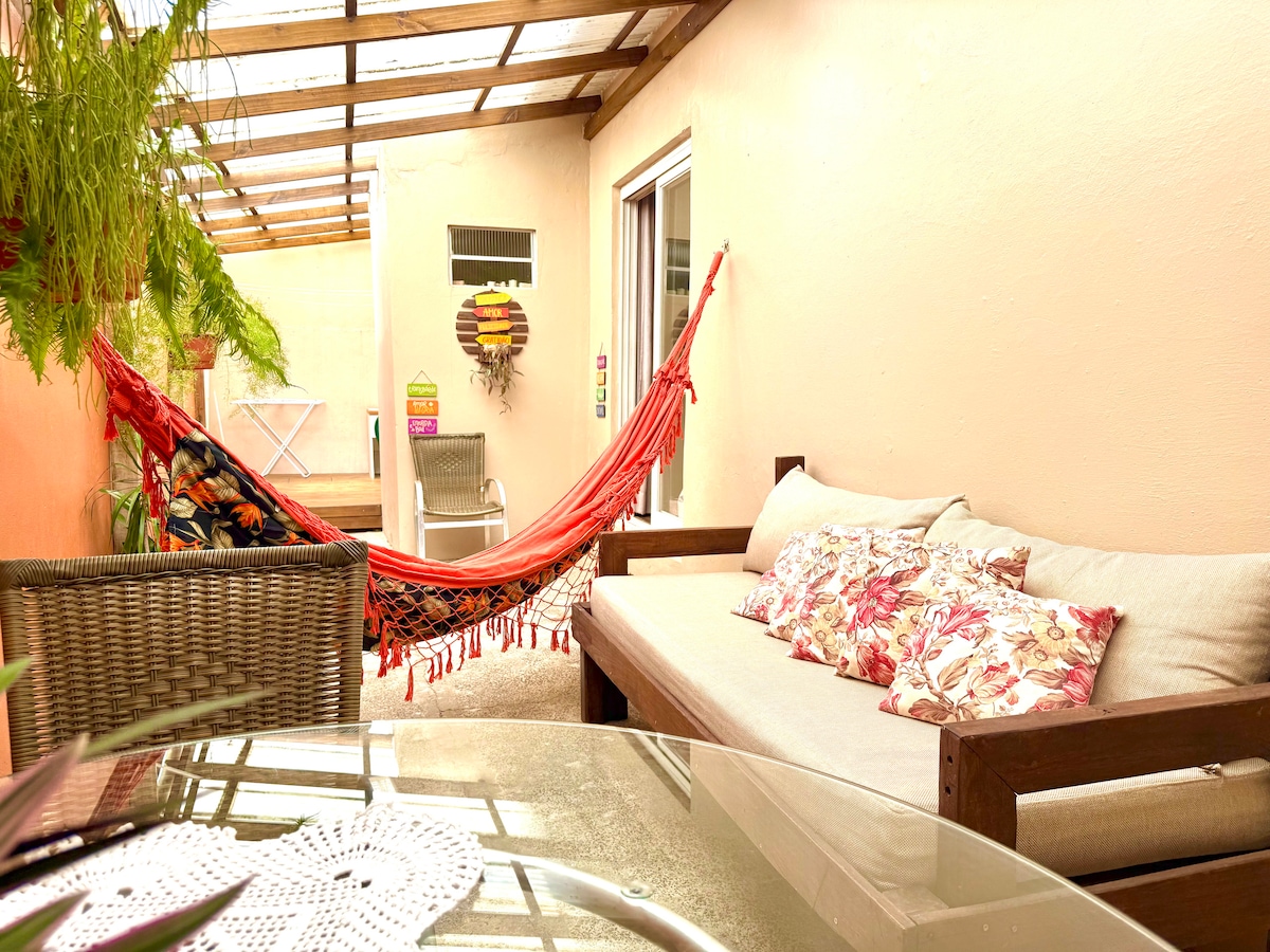 A cozy outdoor patio is featured, showcasing a seating area with a light-colored sofa adorned with decorative cushions. A vibrant hammock is suspended nearby under a shaded pergola, surrounded by leafy plants. A glass-topped table offers a suitable space for relaxation.
