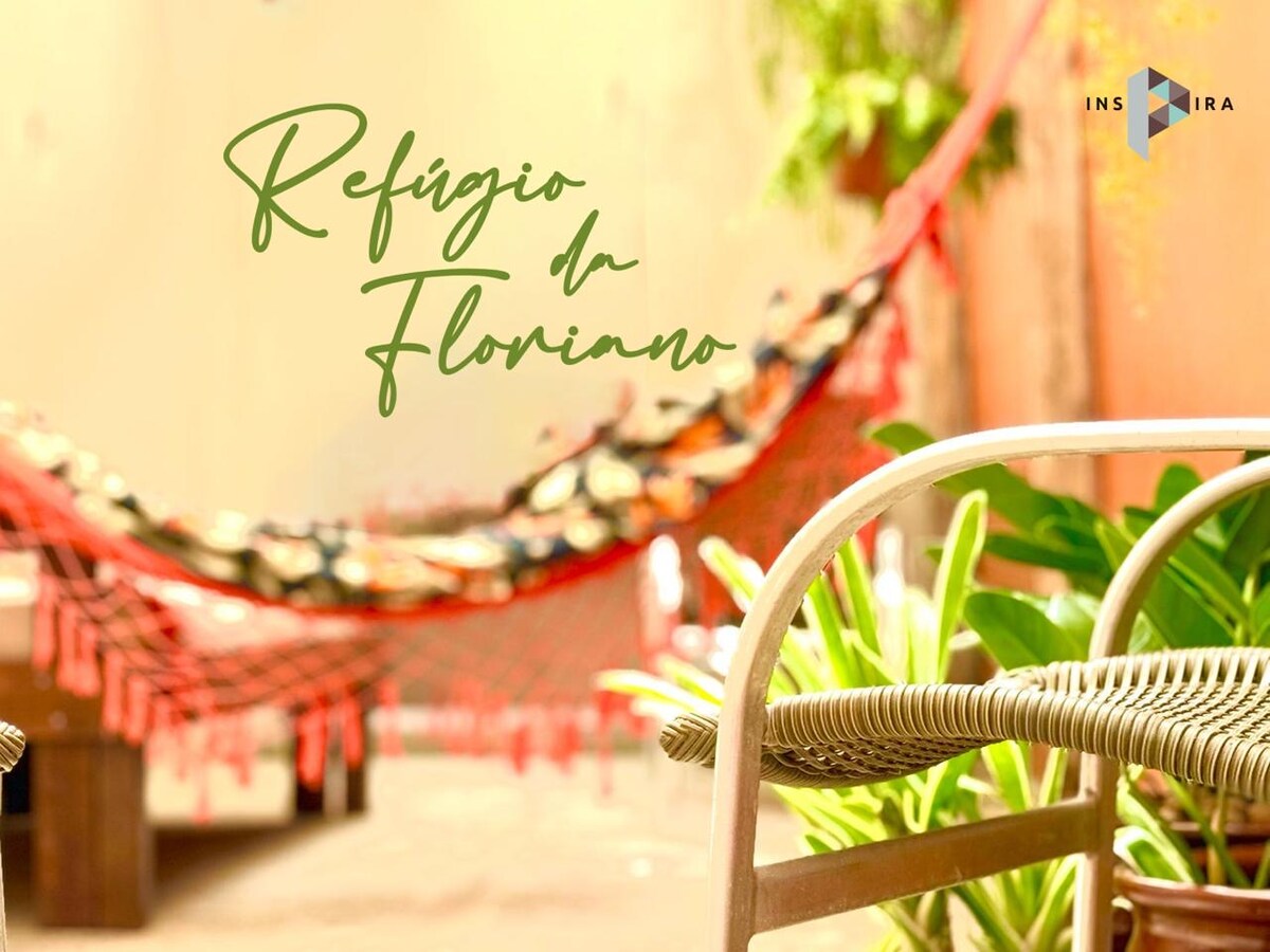 A cozy outdoor space features a vibrant hammock suspended between two walls. Lush green plants are positioned nearby, and a woven chair is seen in the foreground. Natural light enhances the warm atmosphere of this inviting patio.