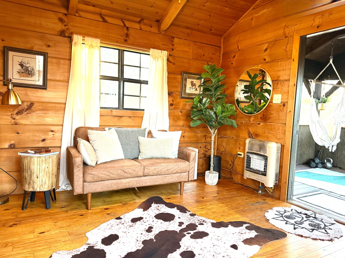 A cozy living area is presented with warm wood paneling and a wooden floor. A brown sofa with decorative pillows sits adjacent to a round mirror, while a small table complements a potted plant. Natural light filters through white curtains framing a nearby window.