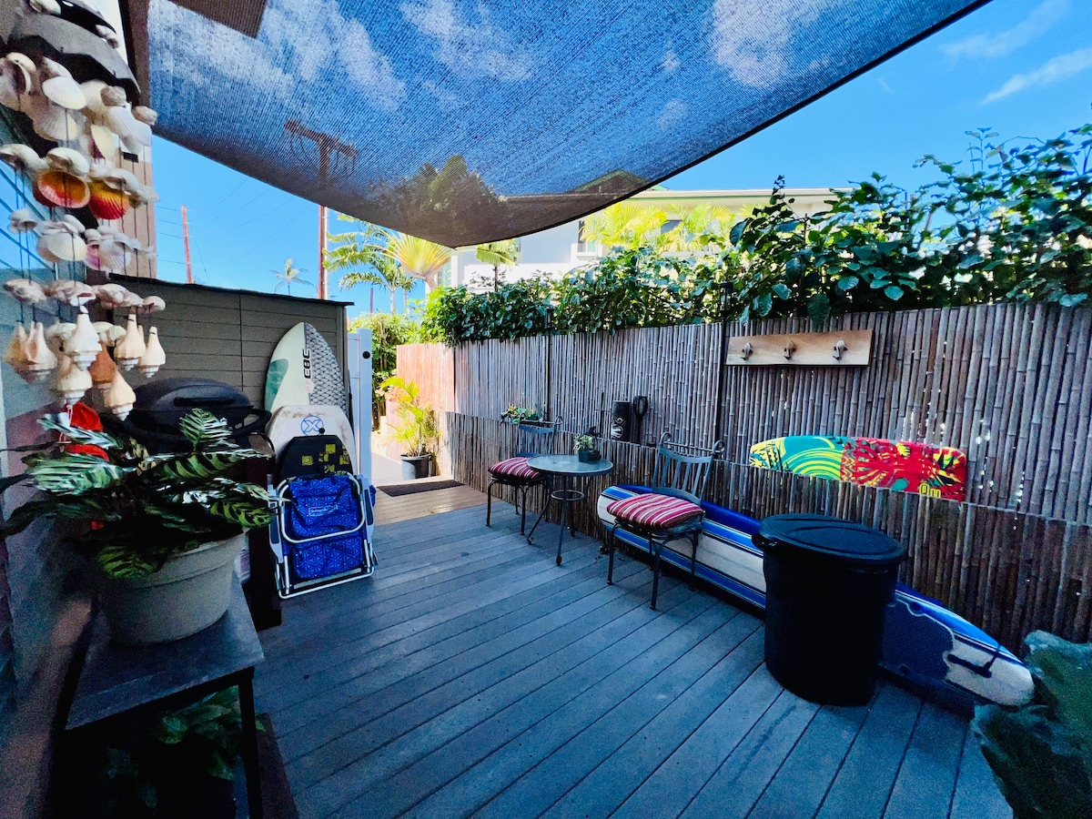 A private rear deck features a shaded area with tropical foliage and bamboo fencing. Two surfboards and beach gear are neatly stored, along with a round table and colorful chairs for seating. A large black container sits in the corner, adding to the functional outdoor space.