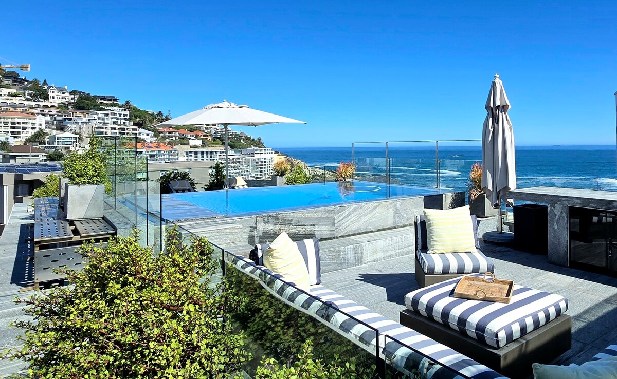 A serene rooftop pool deck showcases a modern infinity pool overlooking the Atlantic Ocean. Comfortable lounge chairs with striped cushions are arranged around the pool, complemented by a shaded umbrella. Lush greenery frames the space, enhancing the scenic coastal views.