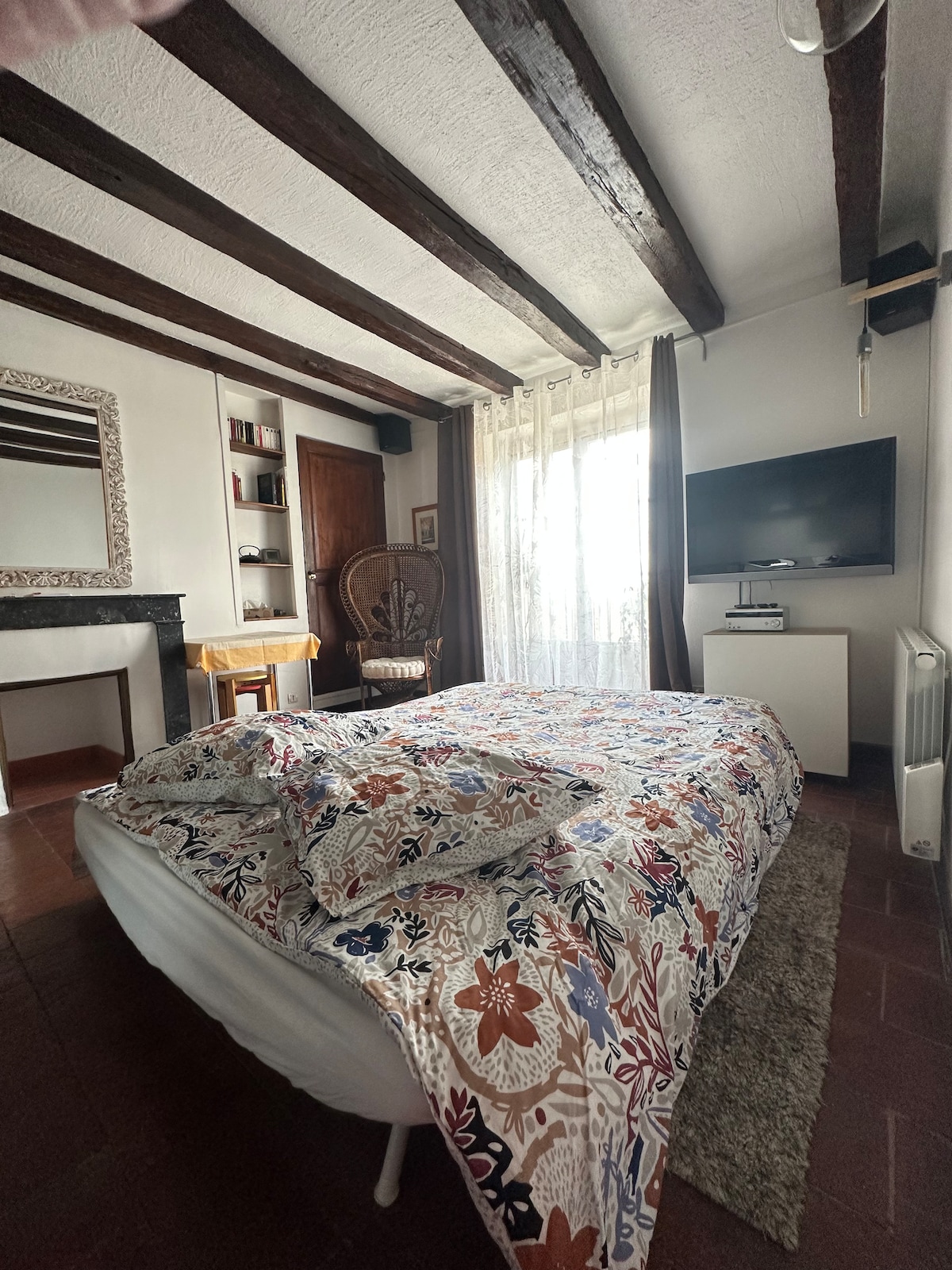 A cozy room features a spacious bed covered with a floral patterned blanket. Natural light filters through sheer curtains, illuminating the hardwood beams above. A wall-mounted TV, small table, and decorative mirror are visible, contributing to a welcoming and functional space.