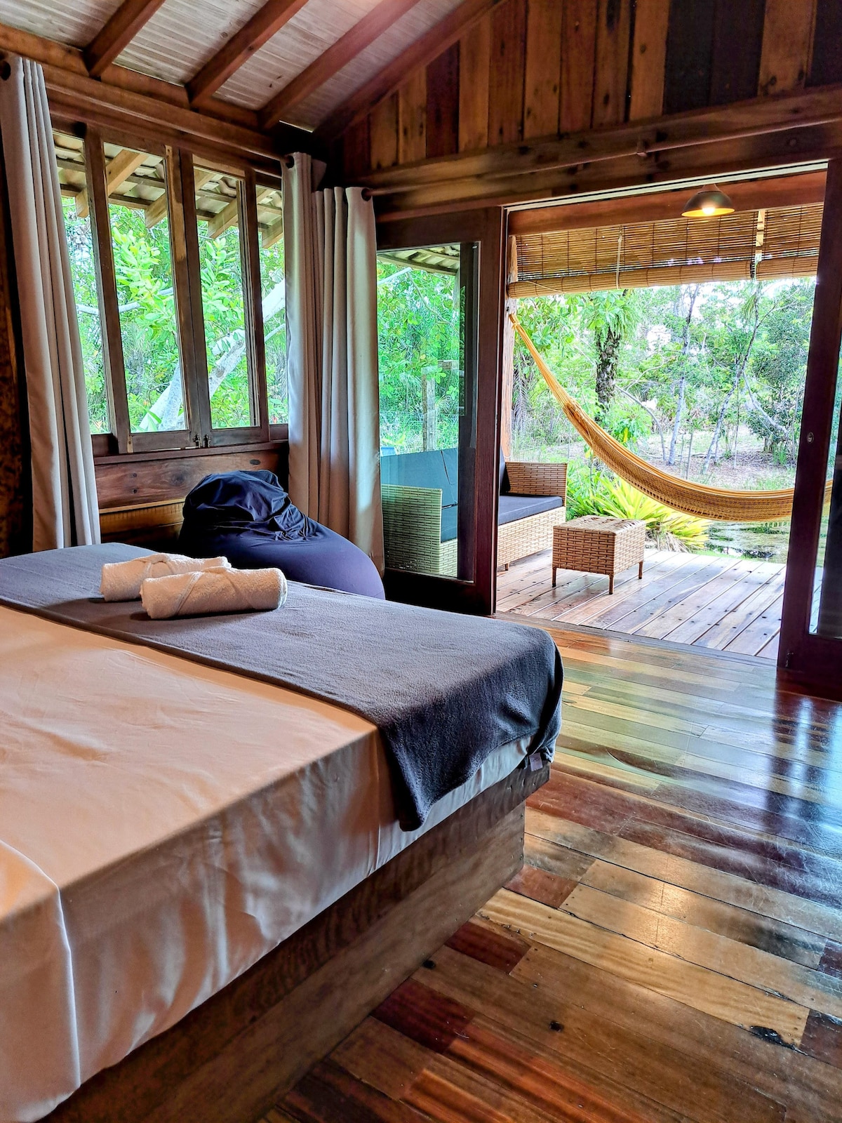 A spacious sleeping area features natural wood flooring and modern furnishings. Large windows allow for views of the outdoor seating area with a hammock. Soft linen is neatly arranged on the bed, complemented by a few rolled towels. The serene setting is enhanced by leafy greenery outside.