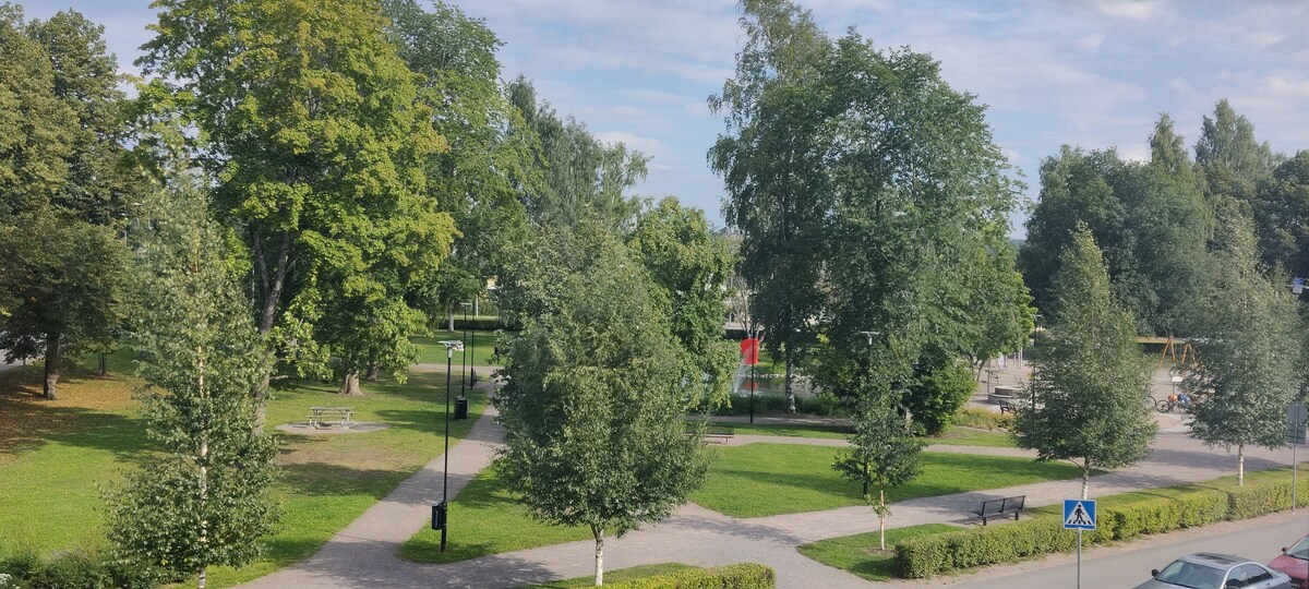 The view features a green park with several trees and well-maintained walking paths. Benches are visible throughout the space, along with a playground area. Lush grass covers the ground, creating a spacious and serene environment for relaxation and leisure activities.