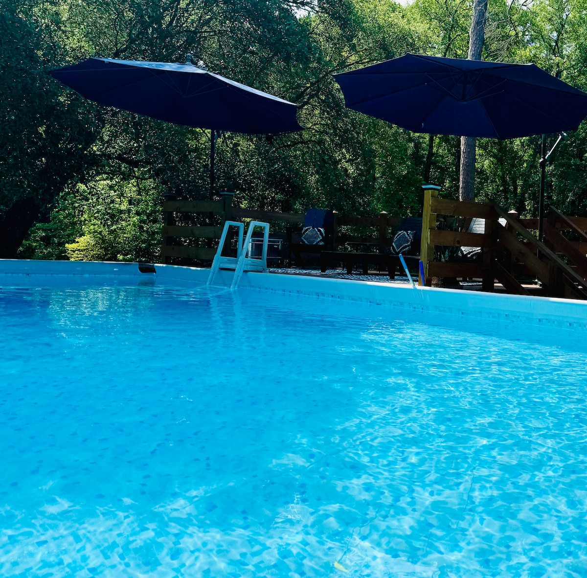 A large above-ground pool is visible, featuring clear blue water and steps leading into it. Two navy umbrellas provide shade along the poolside, where deck seating can be seen against a backdrop of lush green trees.