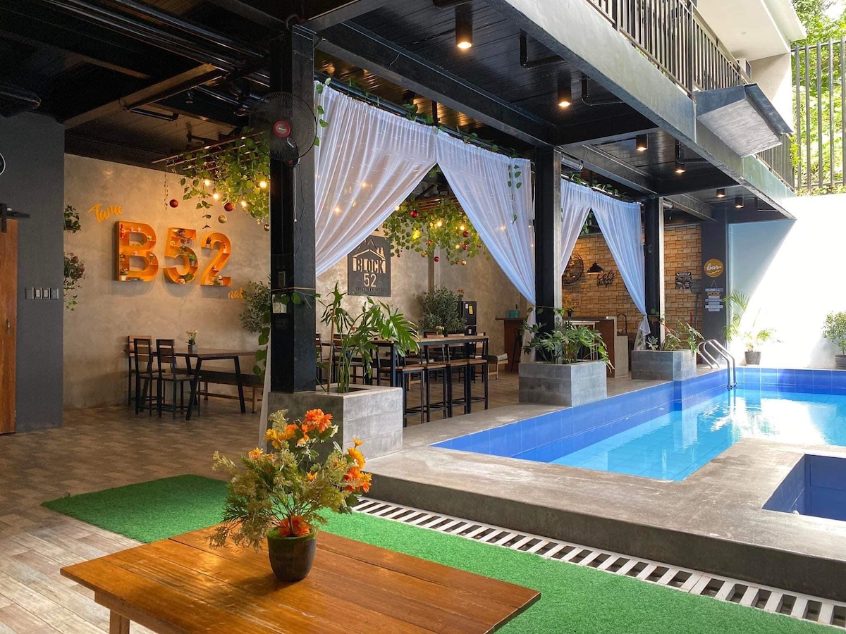 An inviting outdoor area is featured, showcasing a swimming pool adjacent to a dining space. Soft white drapes hang from the ceiling, and the seating is arranged for gatherings. Lush green plants enhance the atmosphere, while a wooden table offers a central focal point.