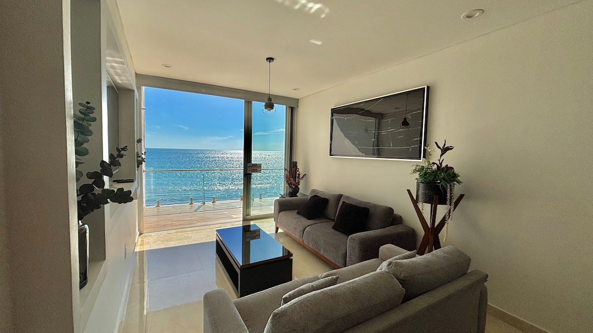 A well-lit living area features two gray sofas positioned around a dark coffee table. Large glass doors offer a view of the ocean and a wooden deck. Decorative plants enhance the space, and modern artwork is displayed on the wall.