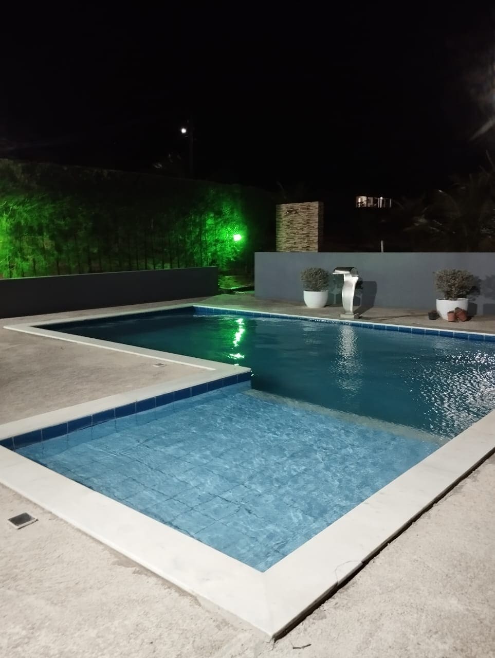 A serene swimming pool is set against a dark backdrop, illuminated by soft green lights. The water reflects a calming blue hue, and potted plants are positioned along the pool's edge, enhancing the tranquil environment.