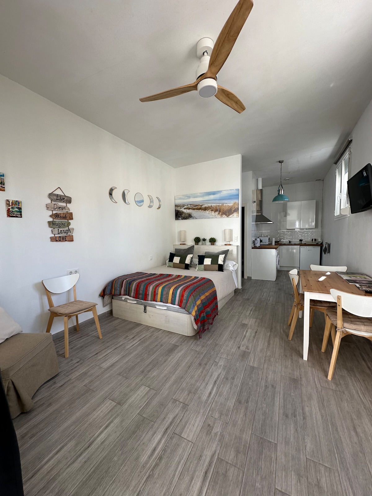 A bright studio space features a cozy sleeping area with a multi-colored striped blanket draped over the bed. A small table and chairs are positioned near the kitchen, creating a functional layout. Natural light enters through the window, enhancing the airy atmosphere.