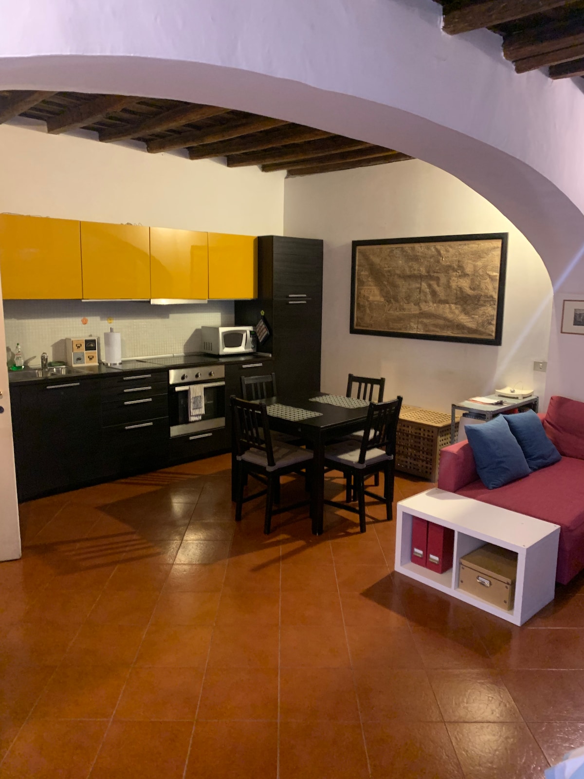 An open living space is showcased, featuring a well-equipped kitchen with yellow cabinetry. A small dining table sits in the center, accompanied by four black chairs. A cozy seating area is visible with a pink sofa and decorative cushions, enhancing the inviting layout.