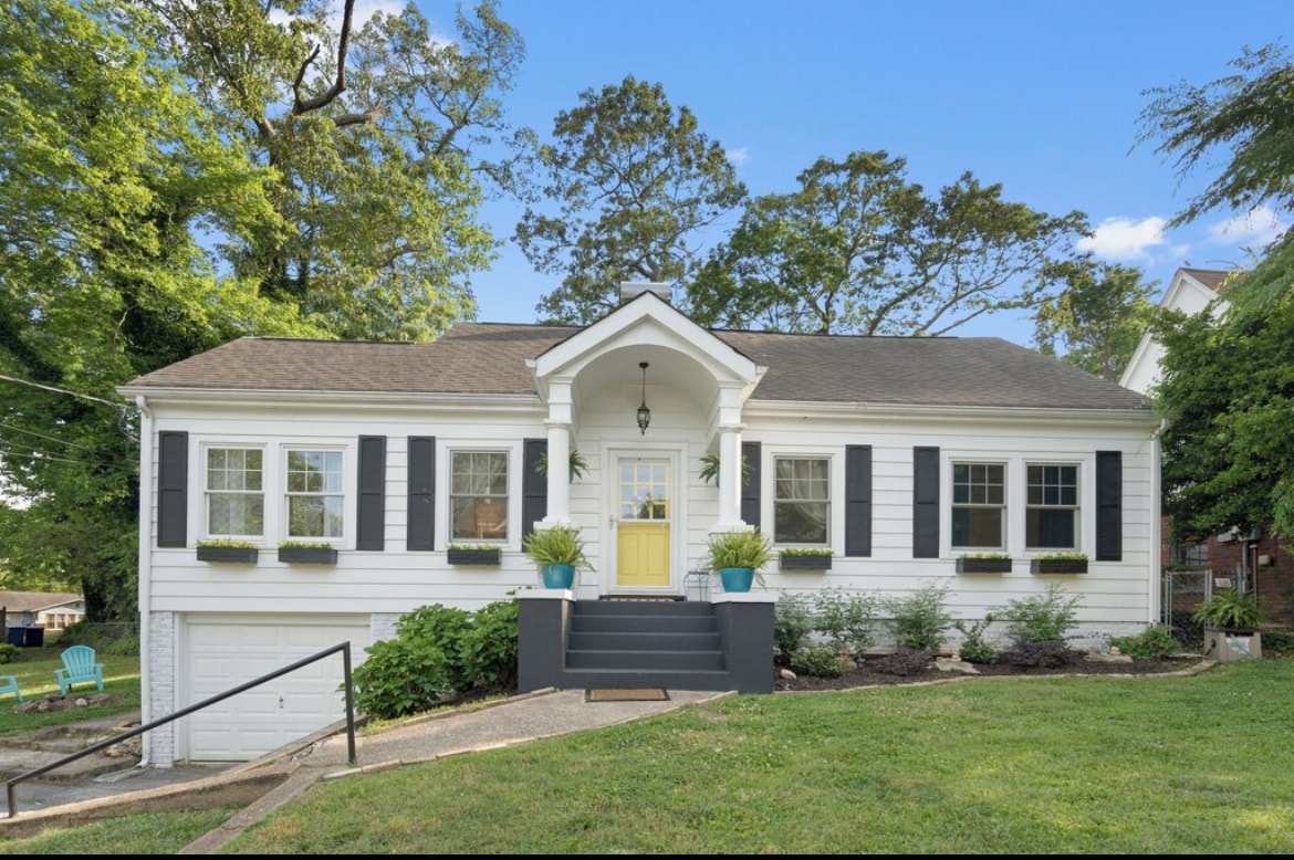 A charming cottage exterior is showcased with white siding and black shutters. A bright yellow door welcomes visitors, while window boxes filled with greenery add a touch of nature. The sloping walkway leads up to the entrance, framed by well-maintained landscaping.