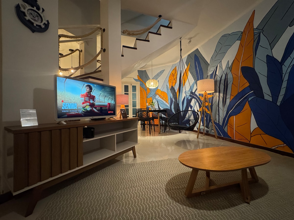 A vibrant living area features a large mural adorned with colorful foliage. A wooden coffee table sits on a textured rug, while a modern television is mounted on a low cabinet. A dining area is visible in the background, accompanied by a decorative light fixture.