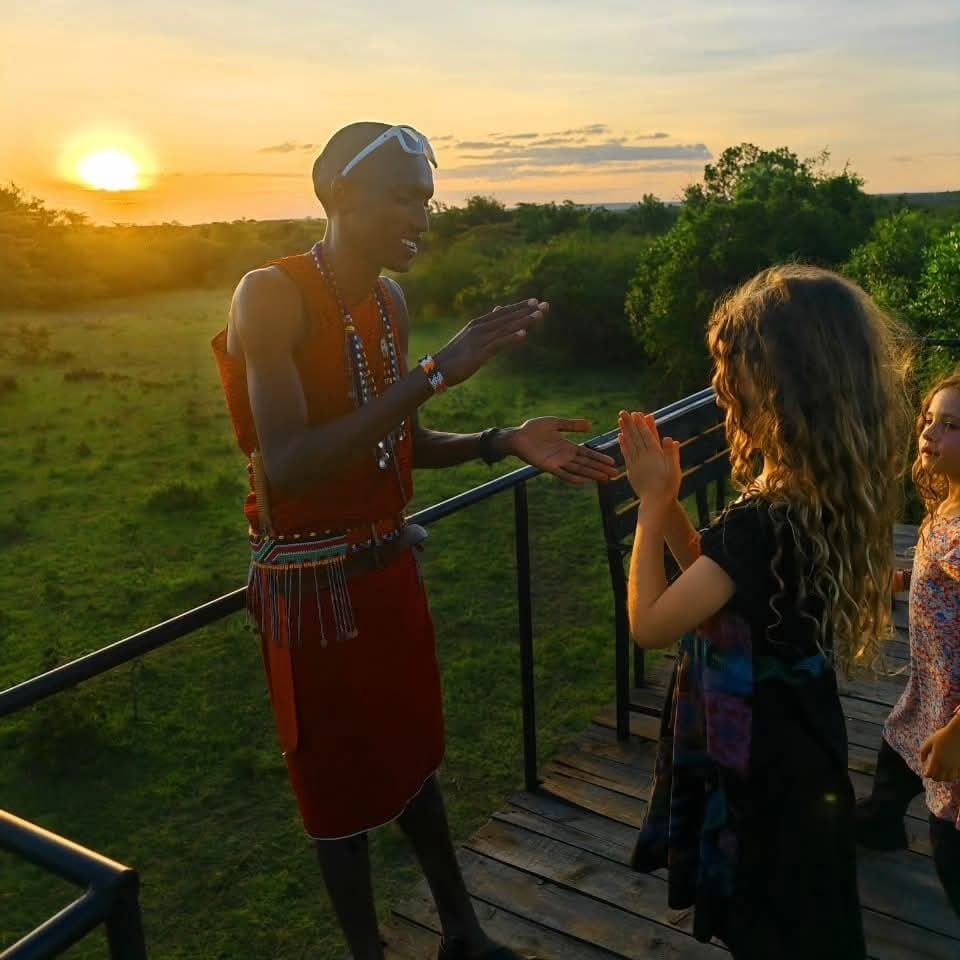 A sunset view captures an expansive landscape, with a Maasai guide engaging with two guests on a balcony. The horizon is painted with warm hues as the sun sets, while lush greenery envelops the scene, representing the serene beauty of the Maasai Mara.