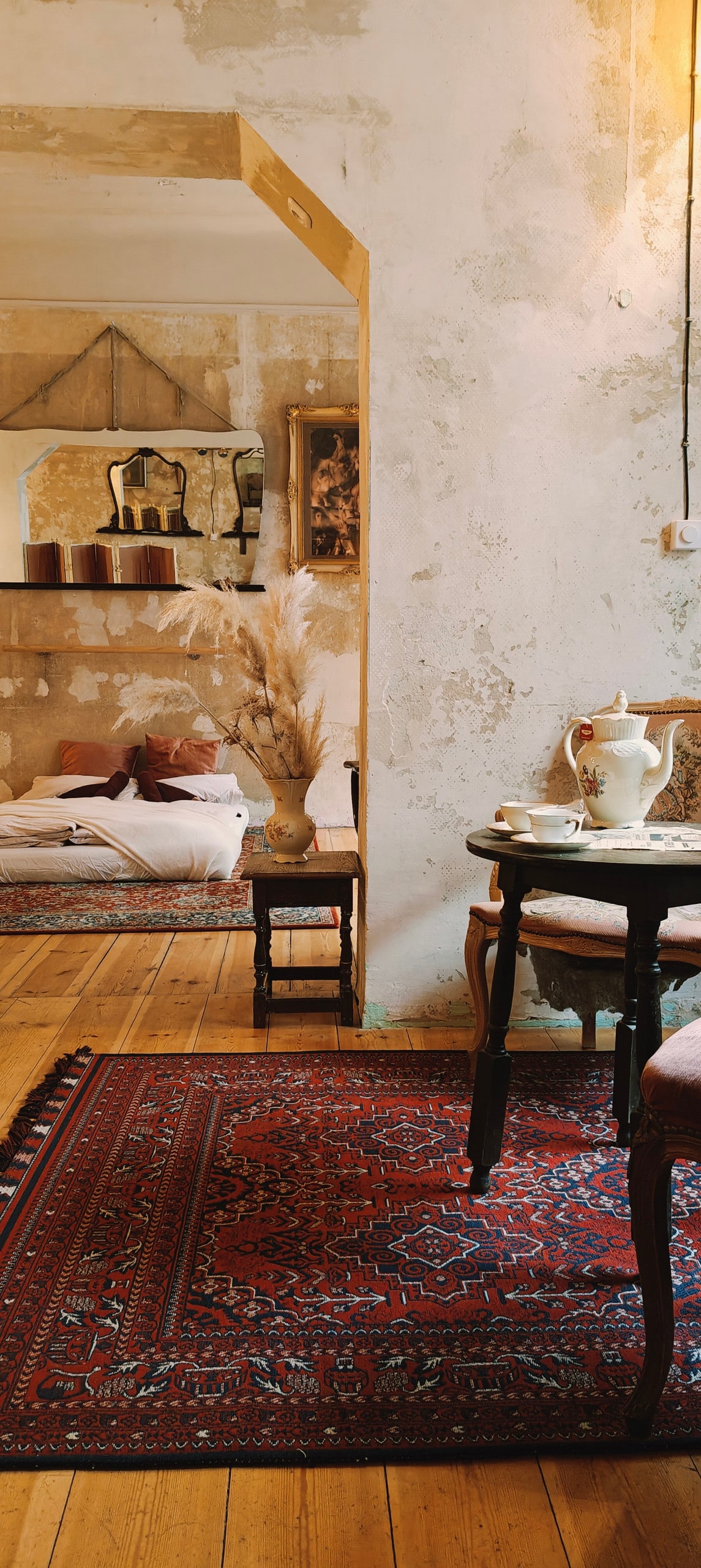 A cozy corner features an ornate rug and a small round table set with an elegant teapot and cups. Soft beige tones dominate the walls, while a glimpse of a bed can be seen through an archway, enhancing the flat's artistic charm.