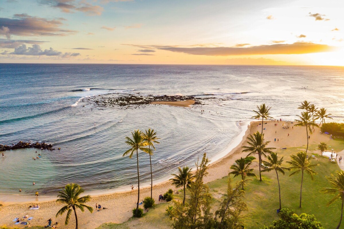 A serene beach scene captures gentle waves lapping on the shore, framed by palm trees. The warm colors of sunset reflect on the water, enhancing the coastal setting. In the distance, beachgoers can be seen enjoying the sandy coastline, while tide pools are visible in the shallow waters.