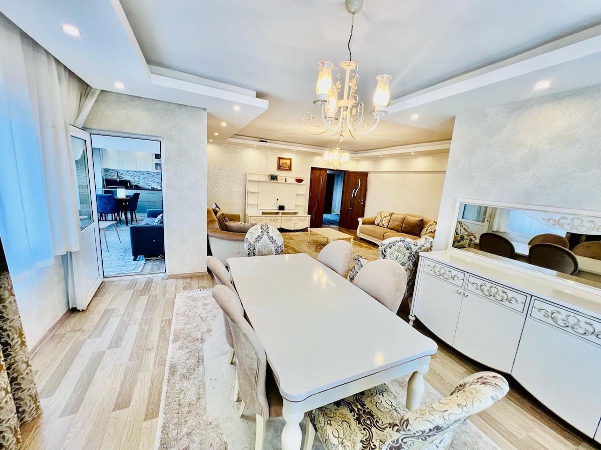 A bright dining area features a large table surrounded by elegant chairs. Soft lighting is provided by a chandelier above, complementing the decorative wall accents. A seating area is visible in the background, with a light-colored couch and a glimpse of outdoor space through sliding glass doors.
