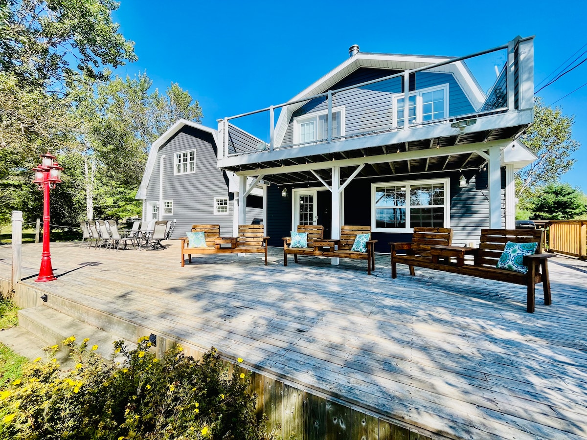 The exterior of the home showcases a spacious deck with comfortable wooden seating and colorful cushions. A large glass door reflects bright sunlight, while the building features contrasting grey siding and a gabled roof amid surrounding trees, enhancing the inviting outdoor environment.