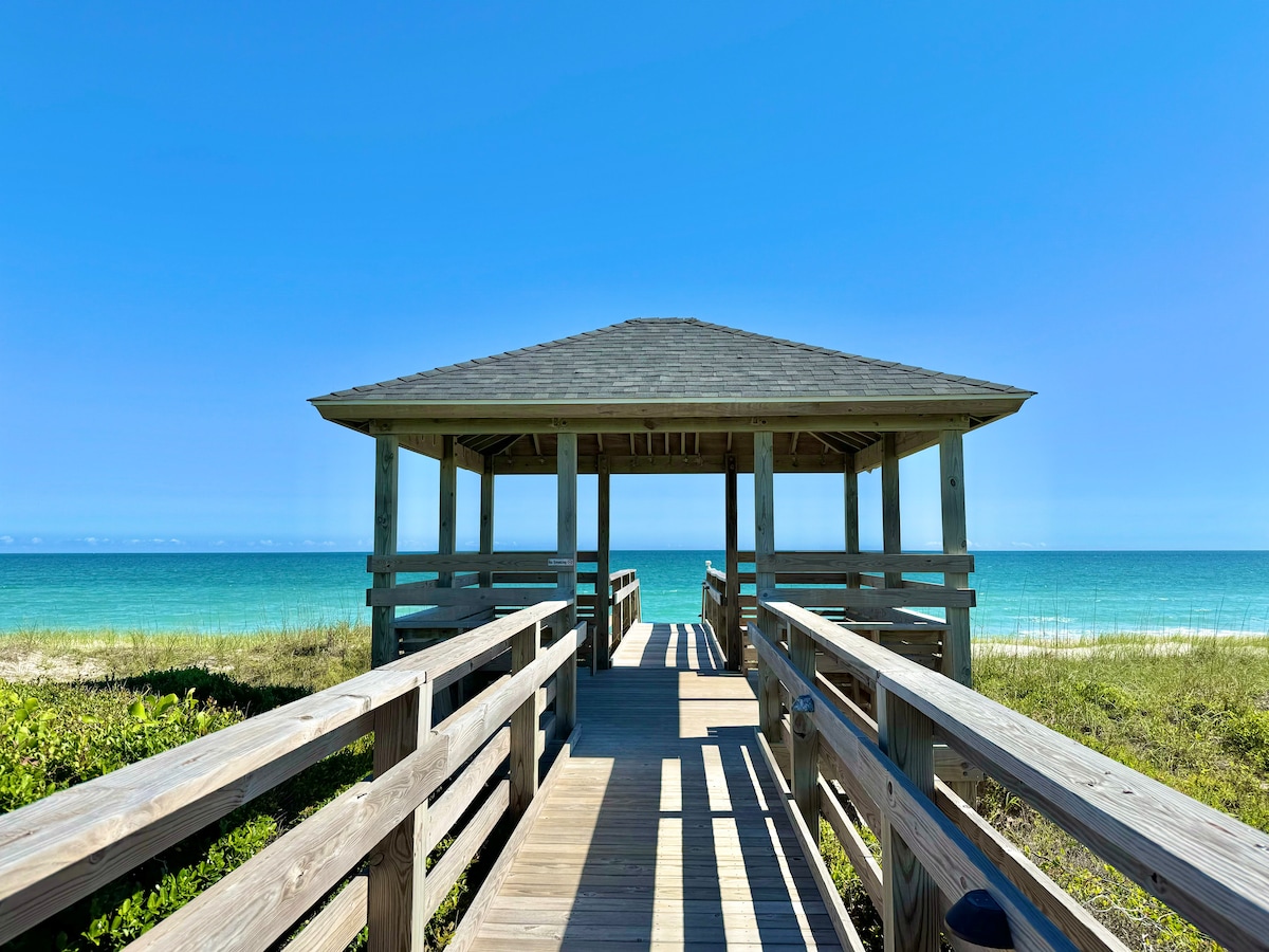 A wooden walkway leads to a spacious gazebo overlooking the ocean. The structure offers a roof and open sides, providing unobstructed views of the serene blue waters and sandy beach. Lush greenery borders the path, enhancing the coastal atmosphere.