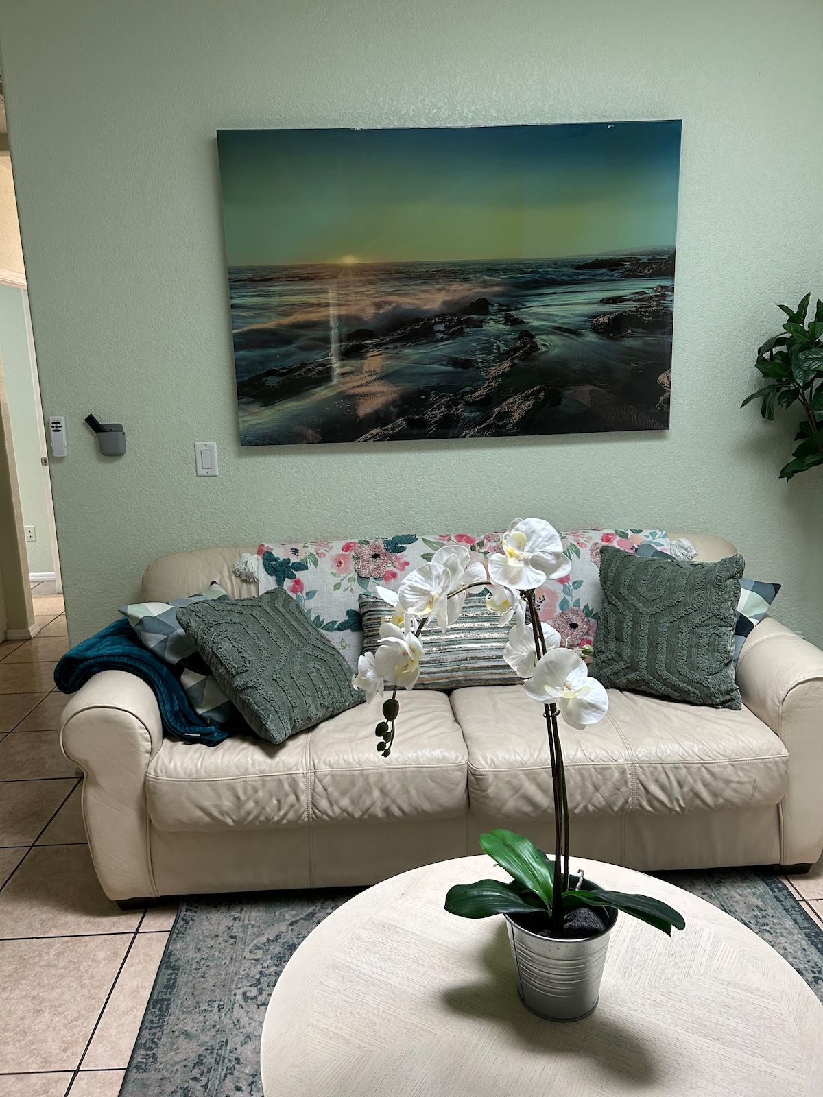 A light-colored couch with a floral throw blanket is positioned in the living area, accompanied by textured pillows in various shades of green. A round coffee table features a potted orchid, and a large artwork depicting a serene seascape is prominently displayed on the wall.
