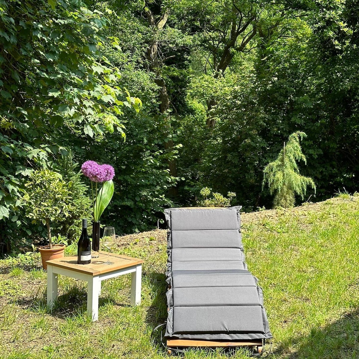 A relaxation area is highlighted by a cushioned lounge chair set on a grassy patch. A small table displays a bottle of wine and two glasses, accompanied by a decorative vase holding a vibrant purple flower. Lush greenery envelops the space, providing a serene backdrop.