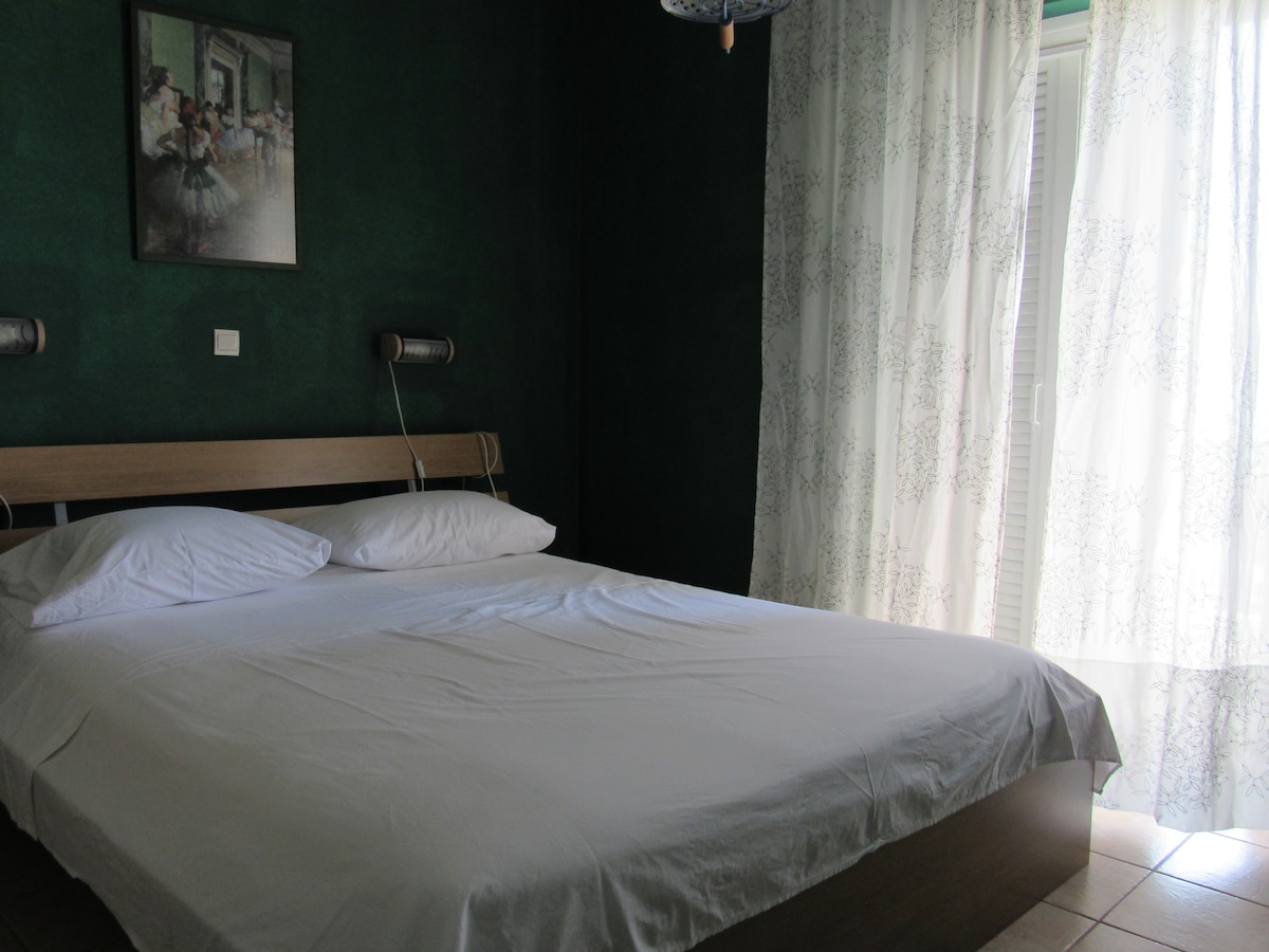 A simple bedroom features a spacious bed with white linens, complemented by two wall-mounted reading lights. Sheer white curtains allow natural light to filter in, and a wall art piece adds a touch of character to the deep green walls.