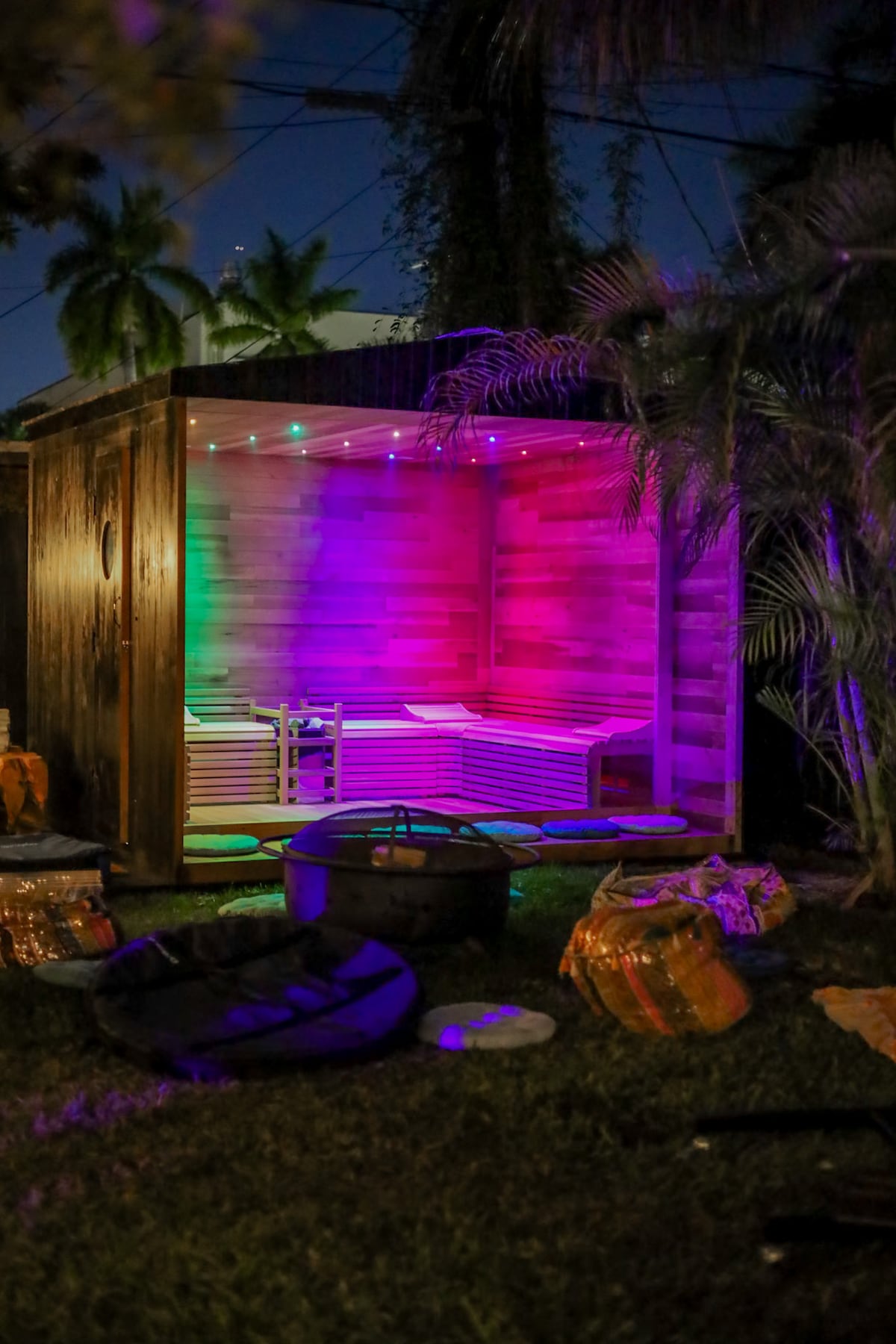 A wooden sauna structure is illuminated with multicolored lights, creating a vibrant atmosphere. Inside, wooden benches are visible, offering seating. The surrounding outdoor area features lush greenery. Various items are placed nearby, adding to the relaxed ambiance of the wellness sanctuary.