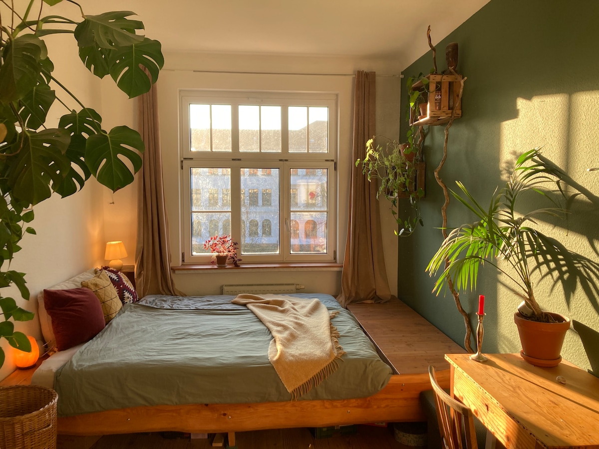 A sunlit bedroom features a spacious bed with a light blanket, positioned near large windows that frame a view of tree branches. Green plants adorn the room, complemented by warm lighting from a bedside lamp. A small wooden table provides a cozy workspace.