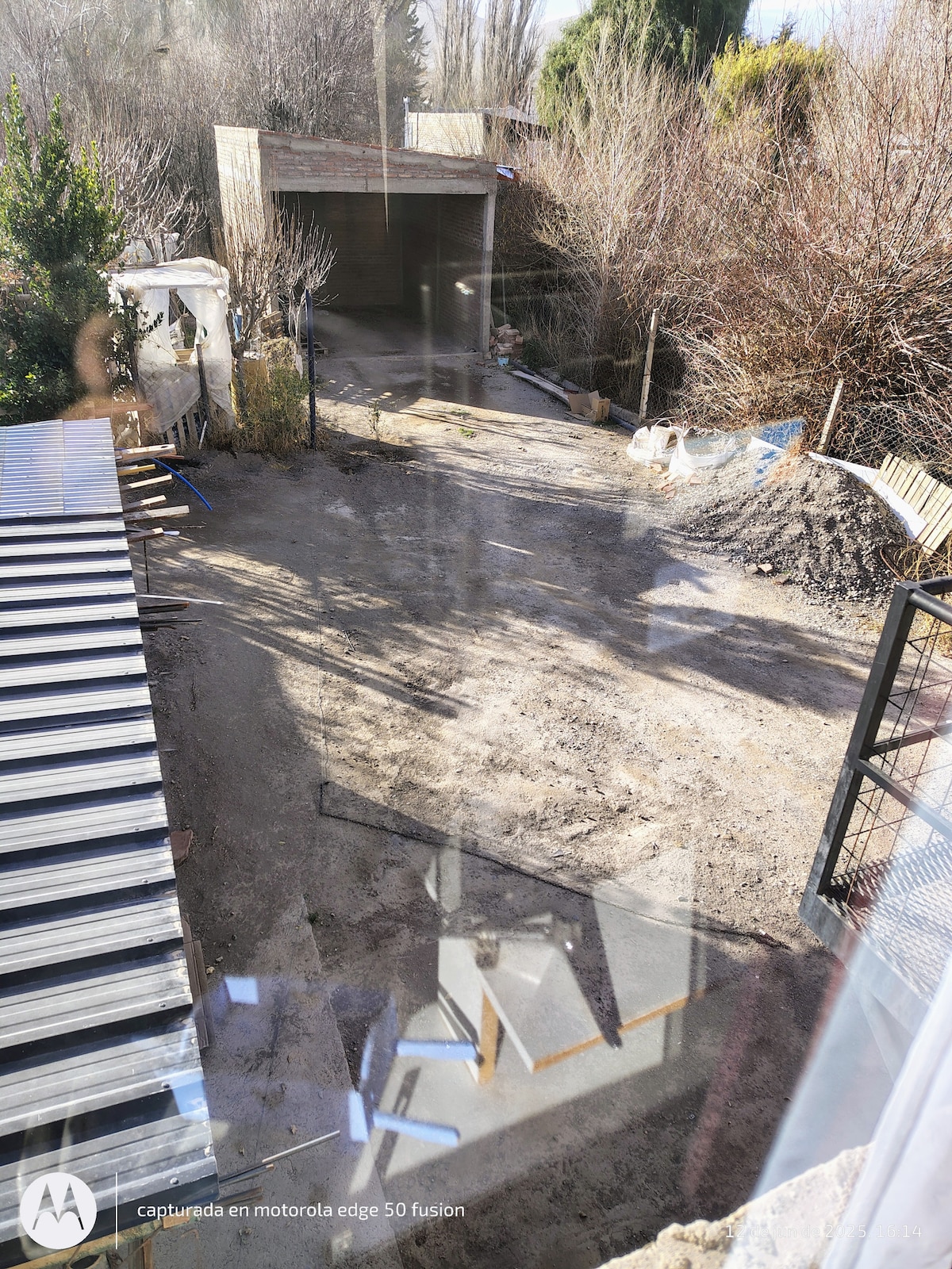 An outdoor area is visible, featuring a gravel surface leading to a shaded parking space. Nearby, a structure with a sloped roof is partially seen, along with sparse vegetation and building materials scattered around. The surroundings appear quiet and rustic.
