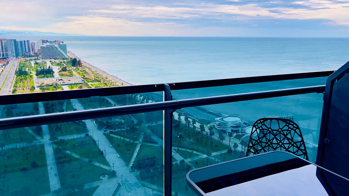 A panoramic view of the coastline is captured from the balcony, highlighting the ocean in varying shades of blue. An outdoor table with a single chair is positioned against the railing, offering a space to enjoy the scenery. Lush green parks are visible below.