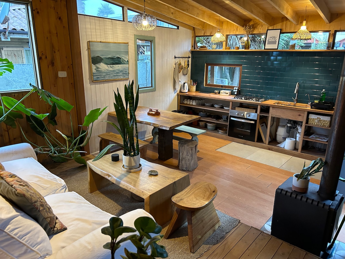 An open living space features natural wood accents and a cozy white sofa. A wooden coffee table sits in front of the sofa, accompanied by plants and a small stool. The kitchen area is showcased with dark green tiles and wooden cabinetry, illuminated by pendant lights.