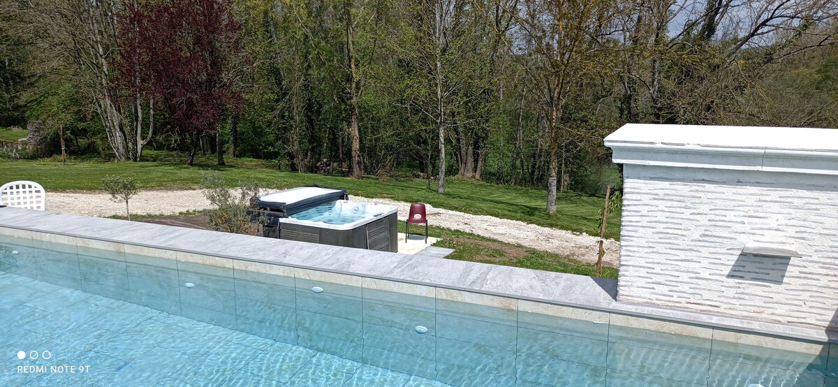 A heated pool glistens under sunlight, with a hot tub situated nearby, surrounded by lush greenery. A pathway is visible in the background, emphasizing the serene and private outdoor setting.