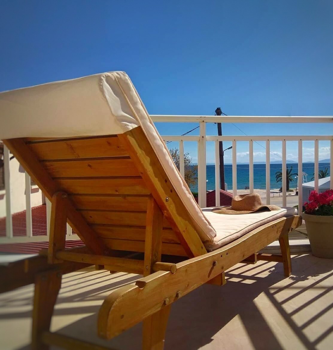 A wooden lounge chair is positioned on a balcony, inviting relaxation with views of the expansive blue Aegean Sea. Bright sunlight bathes the area, highlighting a straw hat resting on the chair's arm. Colorful flowers in a pot add a vibrant touch to the scene.