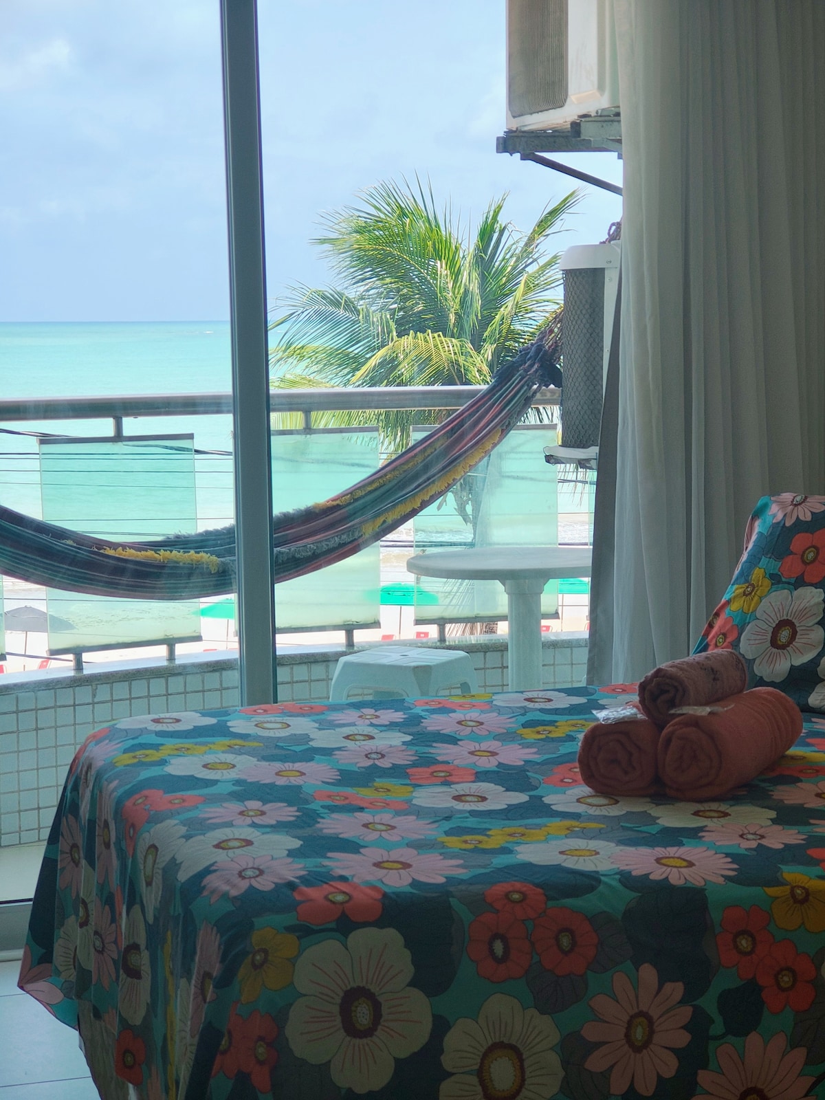 A cozy room is seen with a colorful floral bedspread and rolled towels arranged on the bed. The large glass door leads to a balcony featuring a view of the ocean and palm trees, while a hammock adds a touch of relaxation.