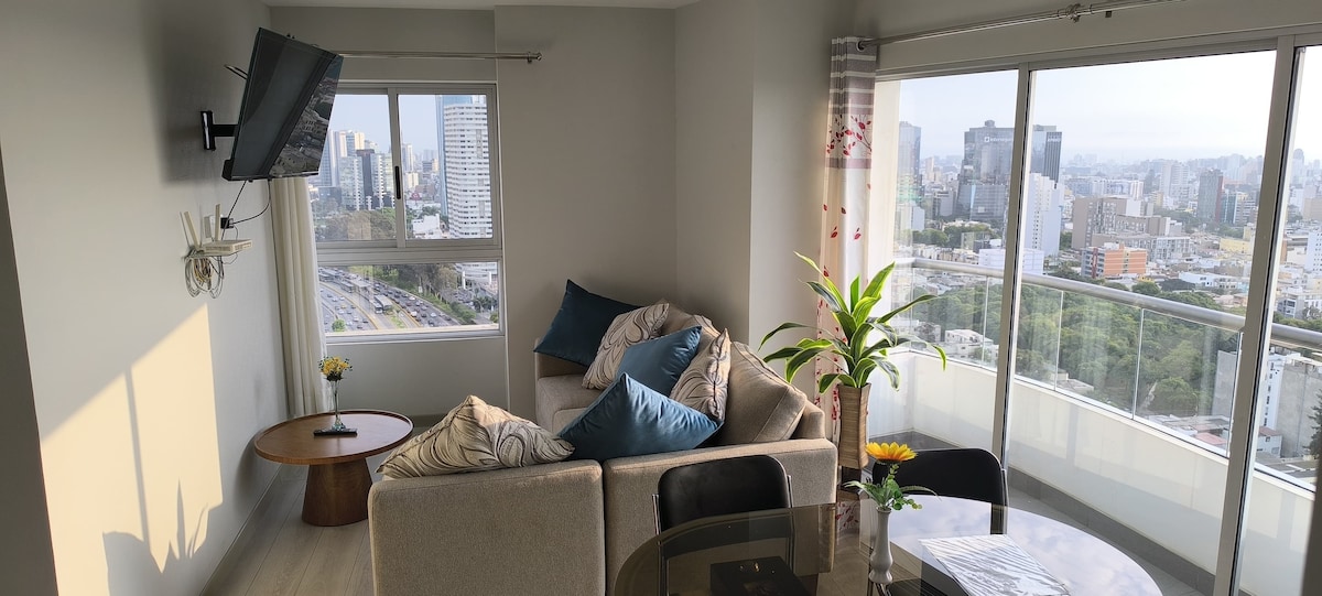 A bright living area features a comfortable sofa adorned with decorative pillows. A wooden coffee table sits nearby, complemented by a vibrant plant. Large windows provide panoramic views of the surrounding cityscape, filling the space with natural light.