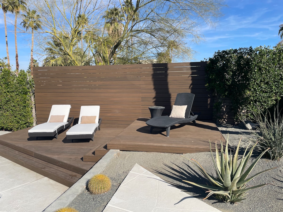 A private outdoor lounge area features two sun loungers and a single chair, arranged on a wooden deck surrounded by desert landscaping. Lush greenery and desert plants provide a serene backdrop against a blue sky, creating an inviting space for relaxation.