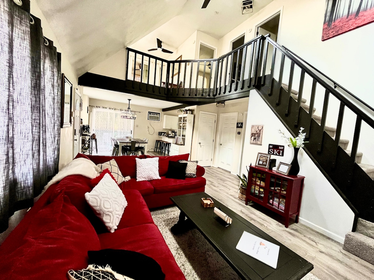 A spacious living area features a large red sectional sofa, cozy decor, and a coffee table atop a textured rug. The open floor plan leads to dining and kitchen areas, while a staircase with black railings ascends to the loft. Natural light filters through large windows, enhancing the inviting ambiance.
