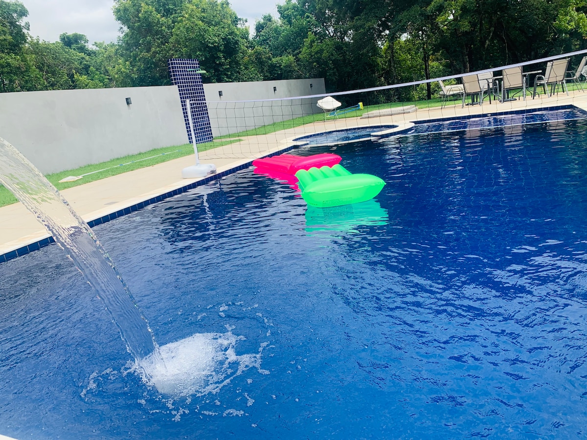 The private swimming pool features a cascading water feature and floats in vibrant pink and green colors. Sun loungers are positioned along the pool's edge, providing a space for relaxation amidst a lush, wooded backdrop.
