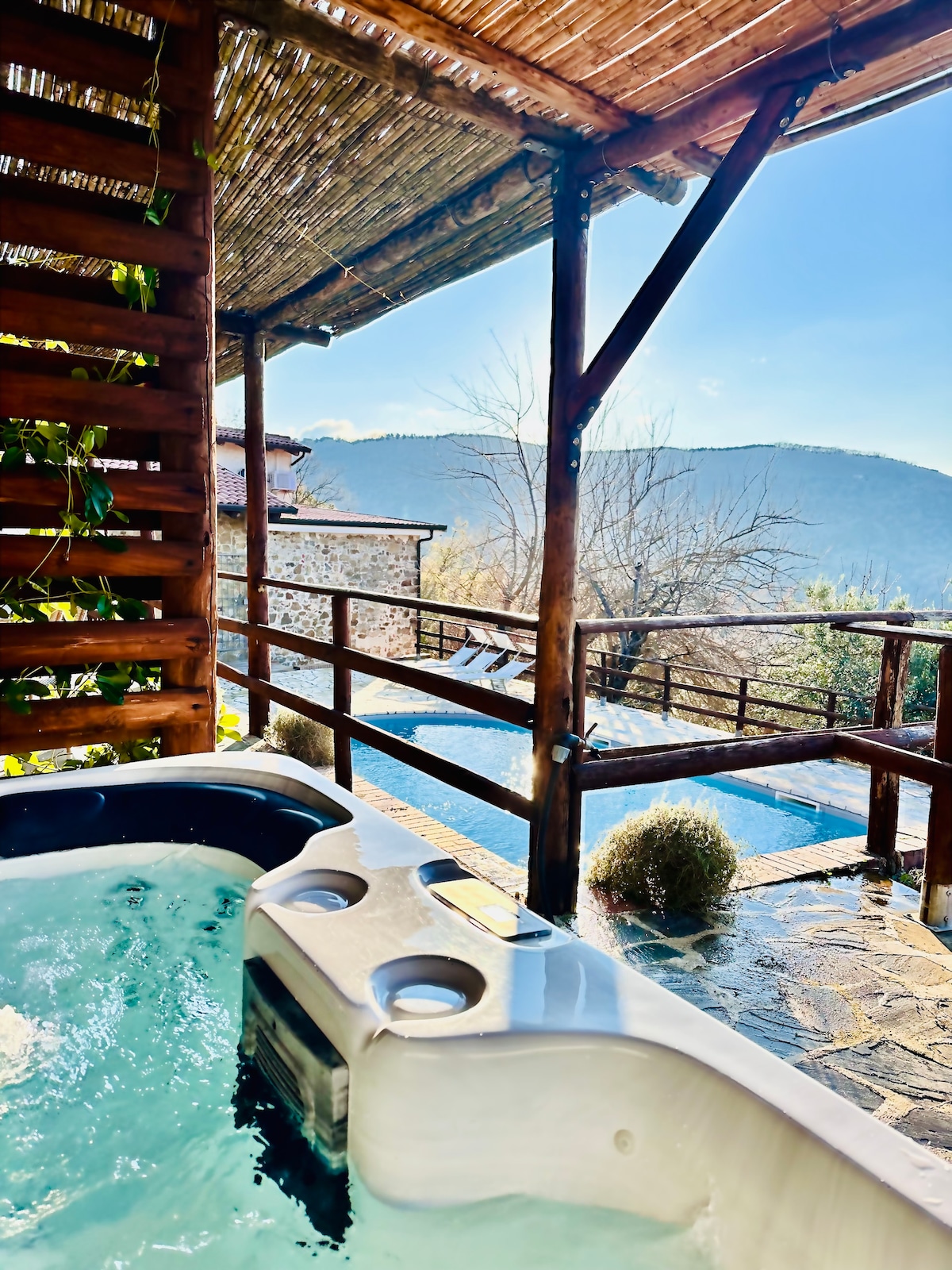 An outdoor jacuzzi is shown alongside wooden decking, surrounded by rustic features. The view in the background reveals distant mountains and a glimpse of a swimming pool. Natural light illuminates the scene, highlighting the tranquil atmosphere.