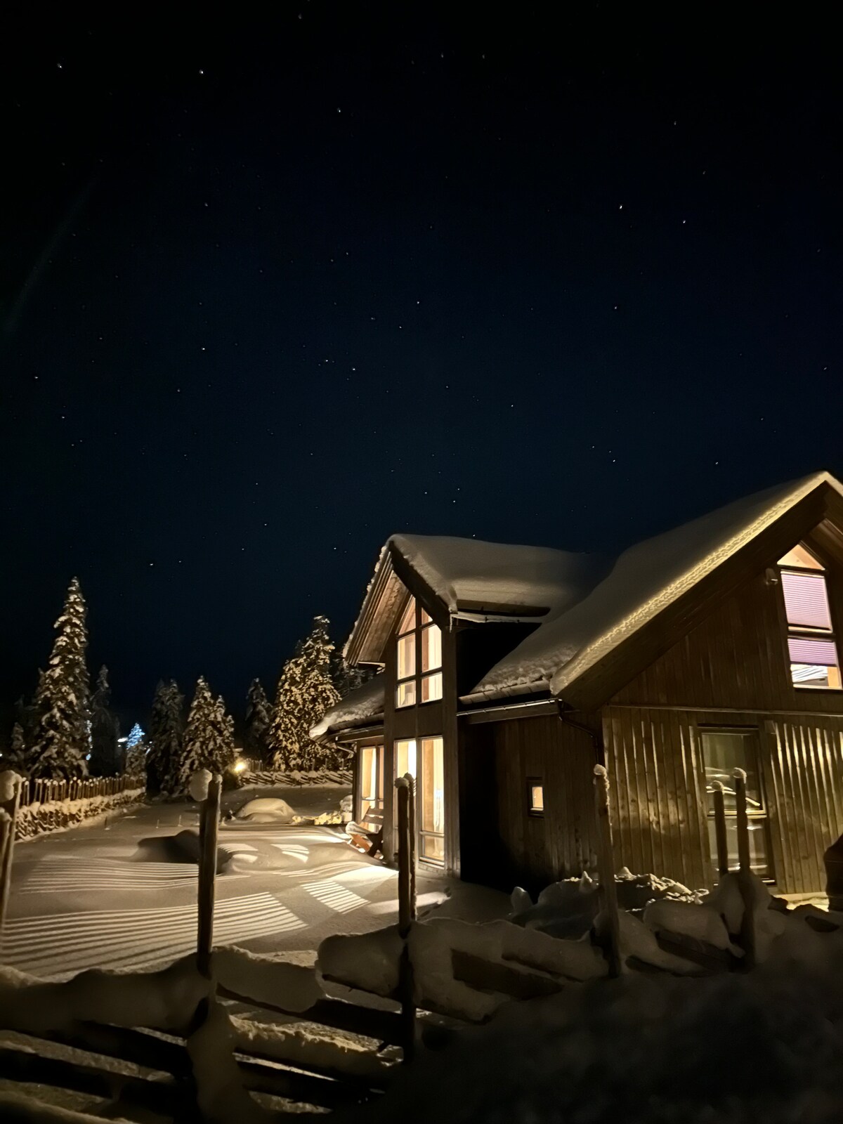 The exterior of the cabin is illuminated under a starlit sky, showcasing a snow-covered roof and surrounding landscape. Warm light spills from the windows, creating a welcoming glow against the snowy backdrop. Trees stand tall nearby, adding to the serene winter scene.
