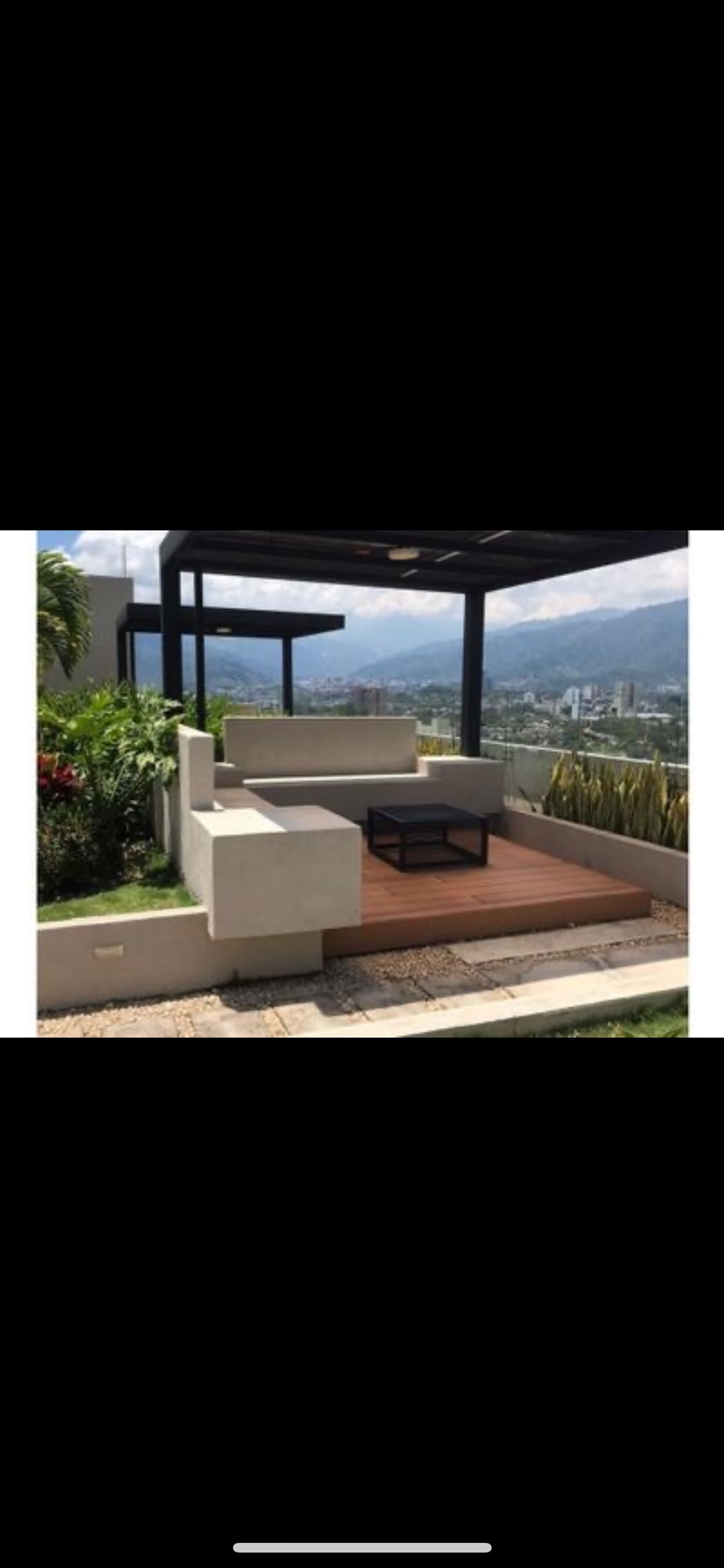 An outdoor seating area is presented, featuring modern concrete furniture arranged around a dark wood table. Surrounded by greenery, the space offers a view of the city and mountains in the background, with a pergola providing partial shade.