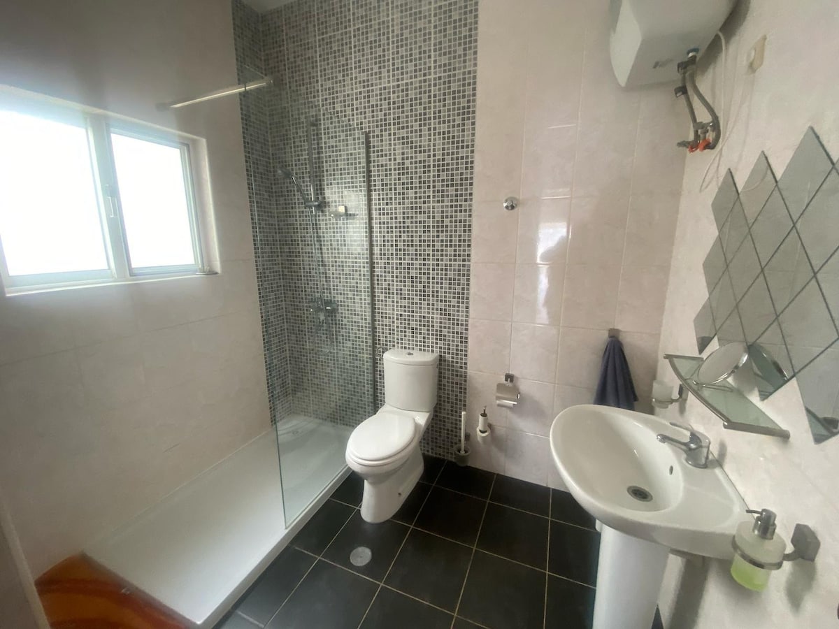 A modern bathroom features a glass-enclosed shower, a white toilet, and a sink with a mirror above. The wall is adorned with gray and black tiles, while natural light enters through a small window, providing a fresh and clean atmosphere.