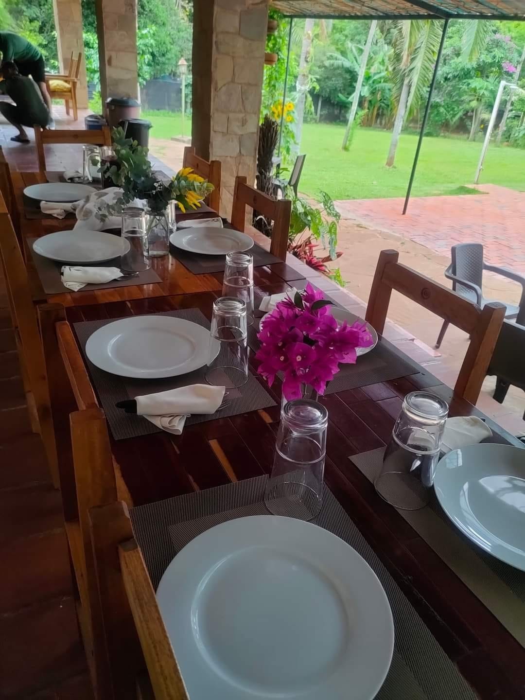 A wooden dining table is set for a meal, featuring empty plates, glasses of water, and neatly folded napkins. A colorful arrangement of flowers sits at the center. The outdoor setting includes greenery and brick flooring, enhancing the inviting atmosphere for gatherings.