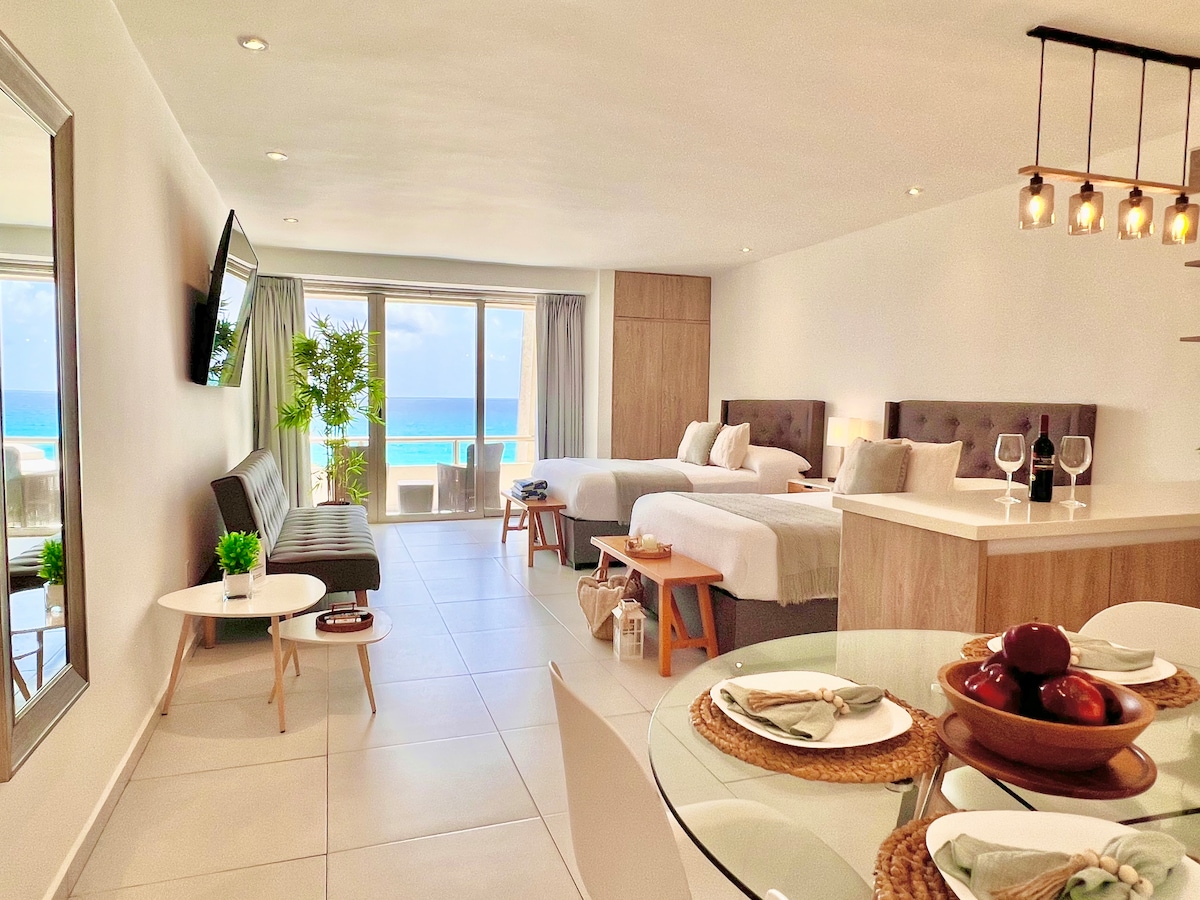 An open-concept studio features two double beds with neutral linens, complemented by a dark gray sofa and a round dining table set for four. Large windows provide views of the Caribbean, and light-colored cabinetry enhances the modern decor throughout the space.