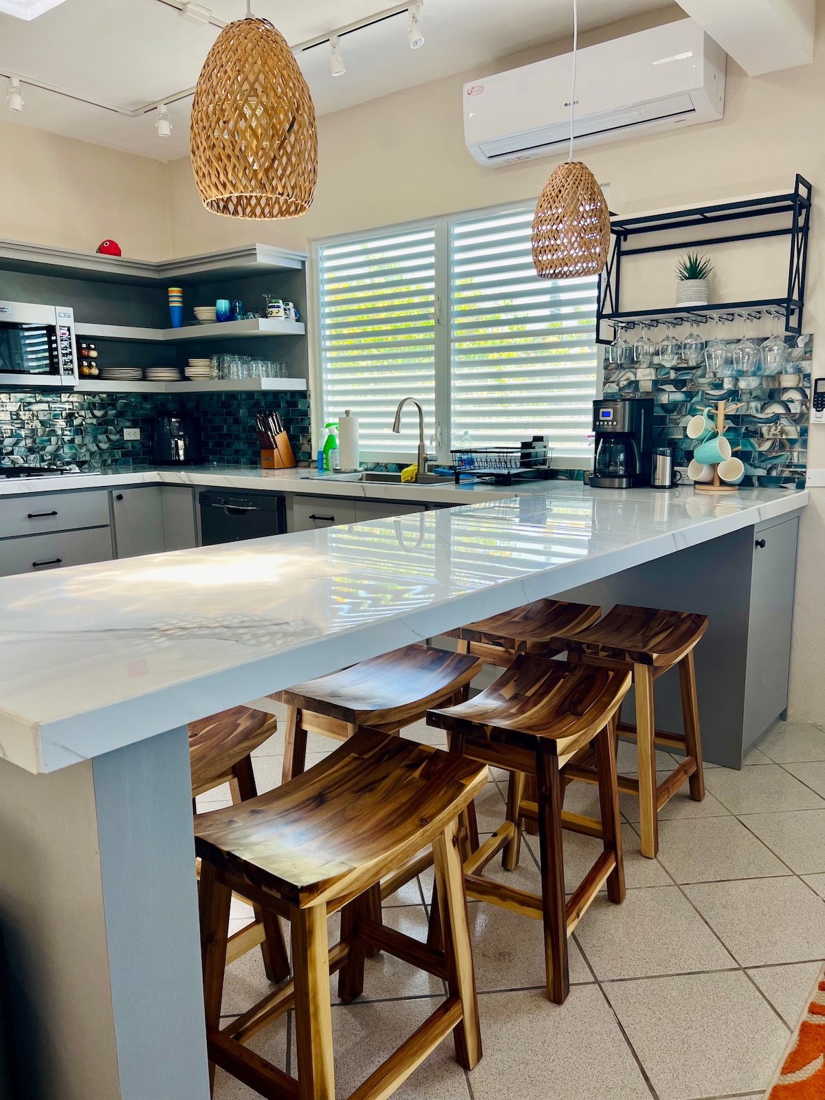 The kitchen features a modern open layout with a spacious island and four wooden stools. Light-colored cabinetry is complemented by a mosaic backsplash and natural light streams through the horizontal window blinds. Essential appliances and utensils are neatly arranged, highlighting a functional cooking space.