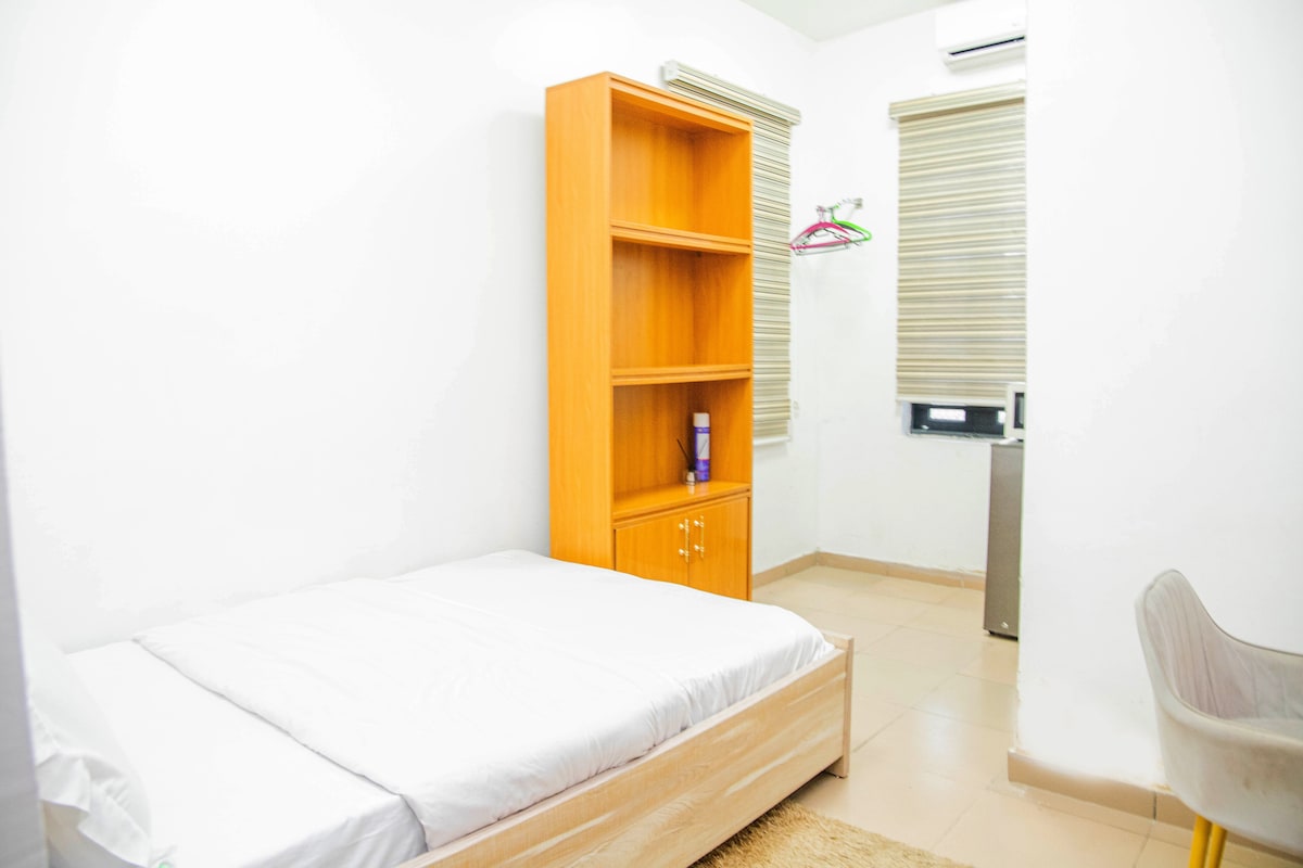 A simple sleeping area features a spacious bed with a white blanket. A wooden shelving unit sits against the wall, complemented by a small chair. Natural light enters through window blinds, and a small refrigerator is visible in the corner.