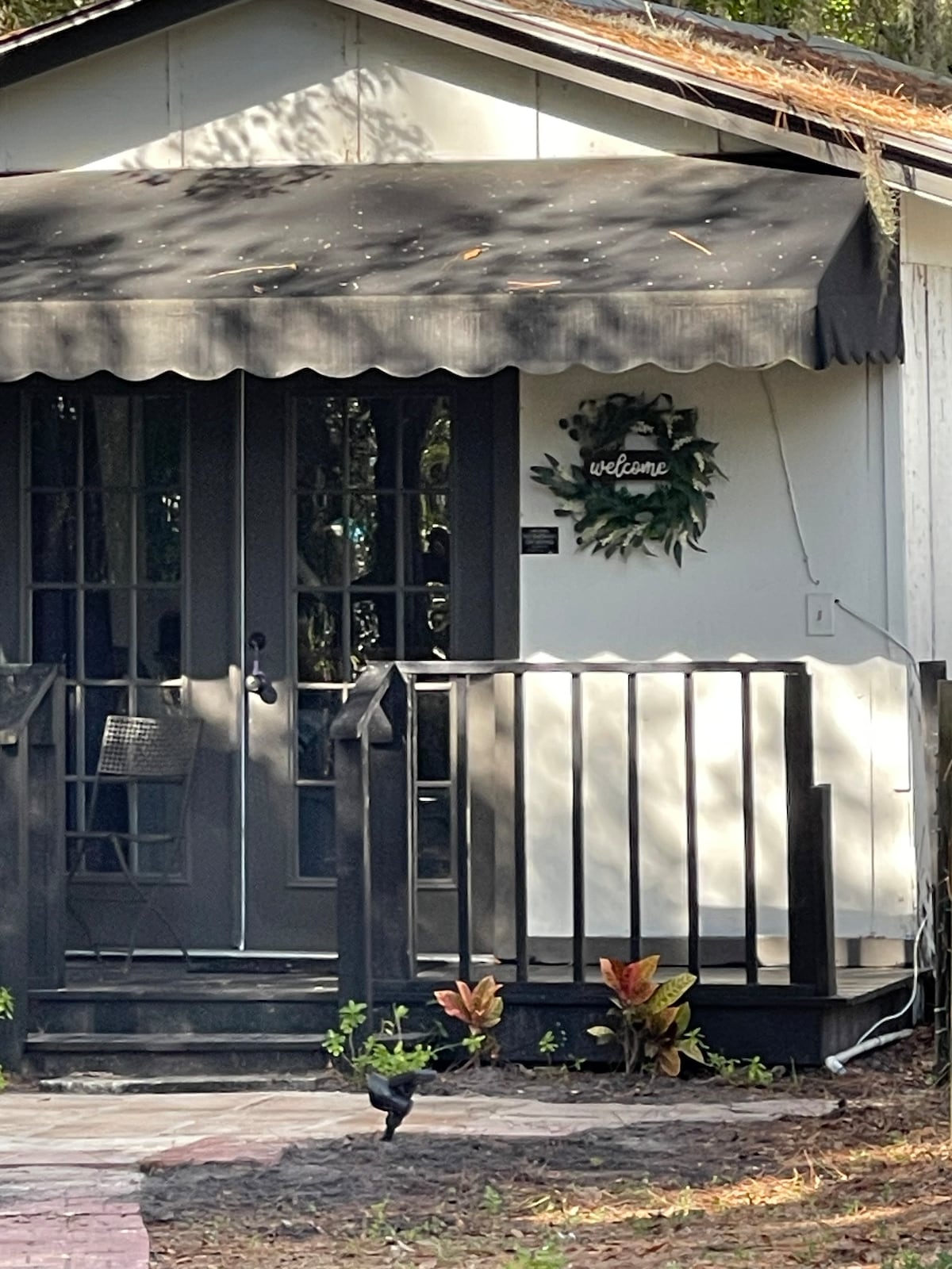 A welcoming entrance is highlighted by a hanging wreath on the door. The porch features a small table and chair, with a stylish awning providing shade. Lush plants are placed around the stairs, enhancing the inviting atmosphere of the cottage.