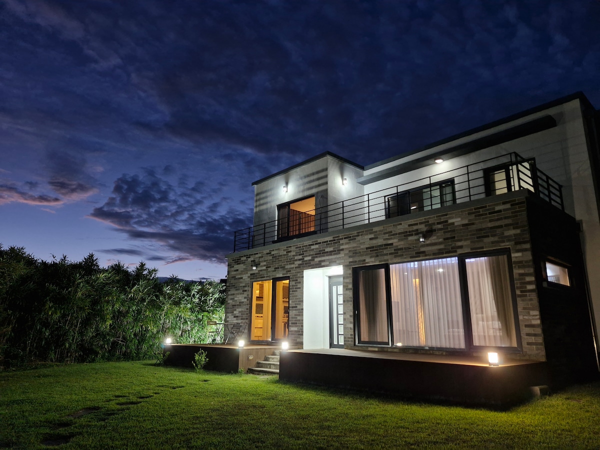 A modern two-story house is featured in the image, illuminated by outdoor lights against a twilight sky. Large windows showcase the inviting interior, while lush greenery is visible around the property, providing a sense of privacy and tranquility.