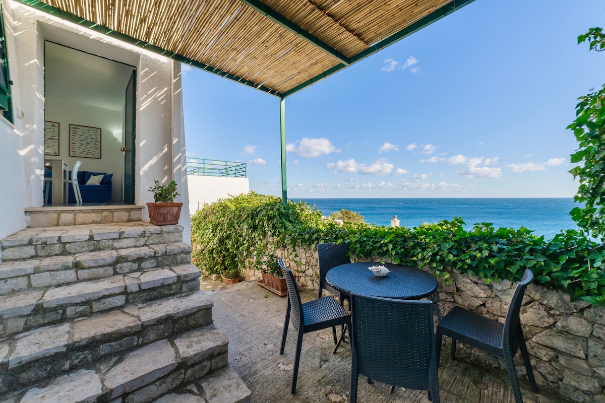 A small outdoor terrace is accessed by stone steps, featuring a round table surrounded by four black chairs. Lush greenery frames the space, with a view of the sea in the background under a clear blue sky.