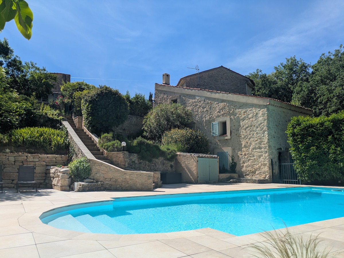 A serene outdoor scene is captured, showcasing a stone house surrounded by lush greenery. A clear swimming pool reflects the sky, with ample space around it for relaxation. Stairs lead from the pool area to the house, enhancing the inviting and tranquil environment.